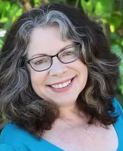 Smiling woman with long wavy hair, gray streaks down the front, wearing a light blue blouse