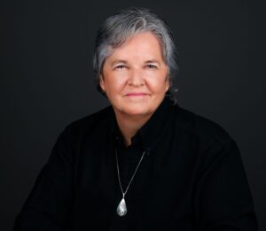 Woman with short gray hair, facing camera, with a dark shirt on against and dark background