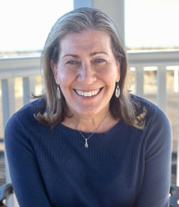 Bright full smile, high cheekbones, a woman with brown sun streaked collar-bone length hair in a dark blue top, sits on a porch