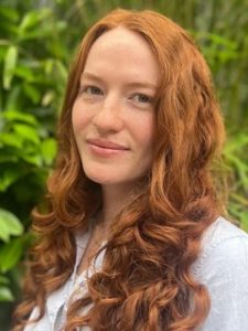 Woman with long red hair which has soft curls cascading over her shoulders. Enigmatic smile.