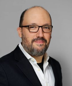 A man with glasses, a short beard, and a receding hairline, wearing a dark blazer over a white collared shirt, smiling slightly, smiling a little mysteriously. He is against a neutral gray background.