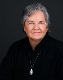 Short salt and pepper haired woman with a calm demeanor and gentl smile, dressed in black, sitting straight looking right through to your soul.